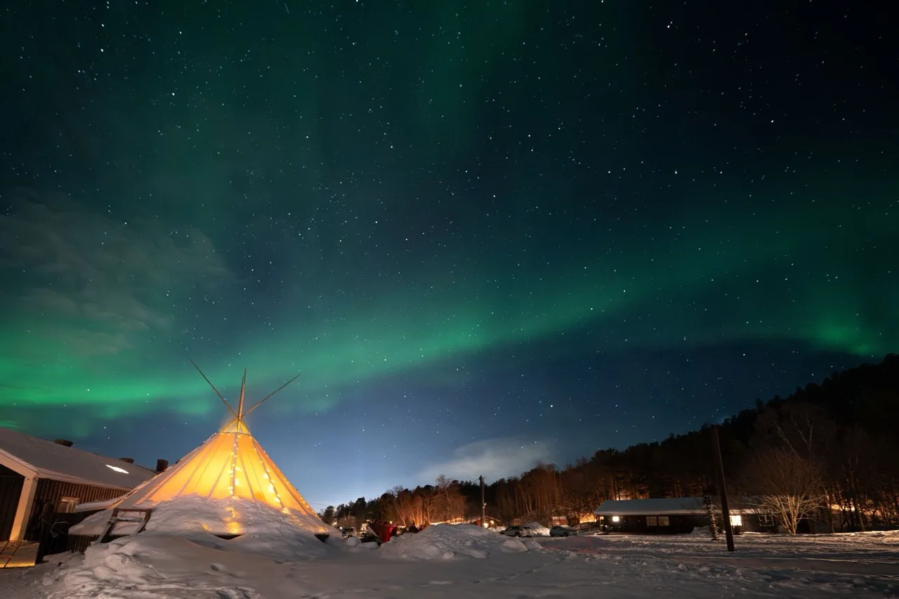Northern lights over a snow-covered property, with a lit tent and lodge buildings
